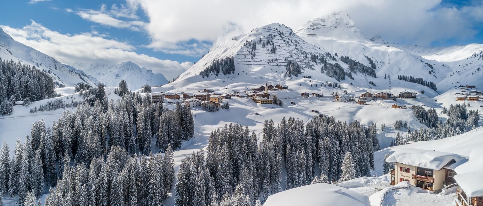 Winterlandschaft mit schneebedeckten Bergen, Tannen und Häusern unter blauem Himmel mit Wolken