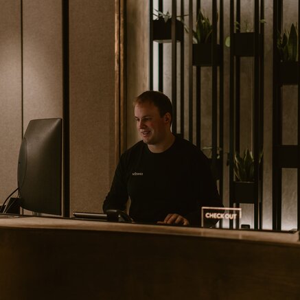 Mann in schwarzem Pullover sitzt lächelnd an einem Schreibtisch mit Computer und "Checkout"-Schild