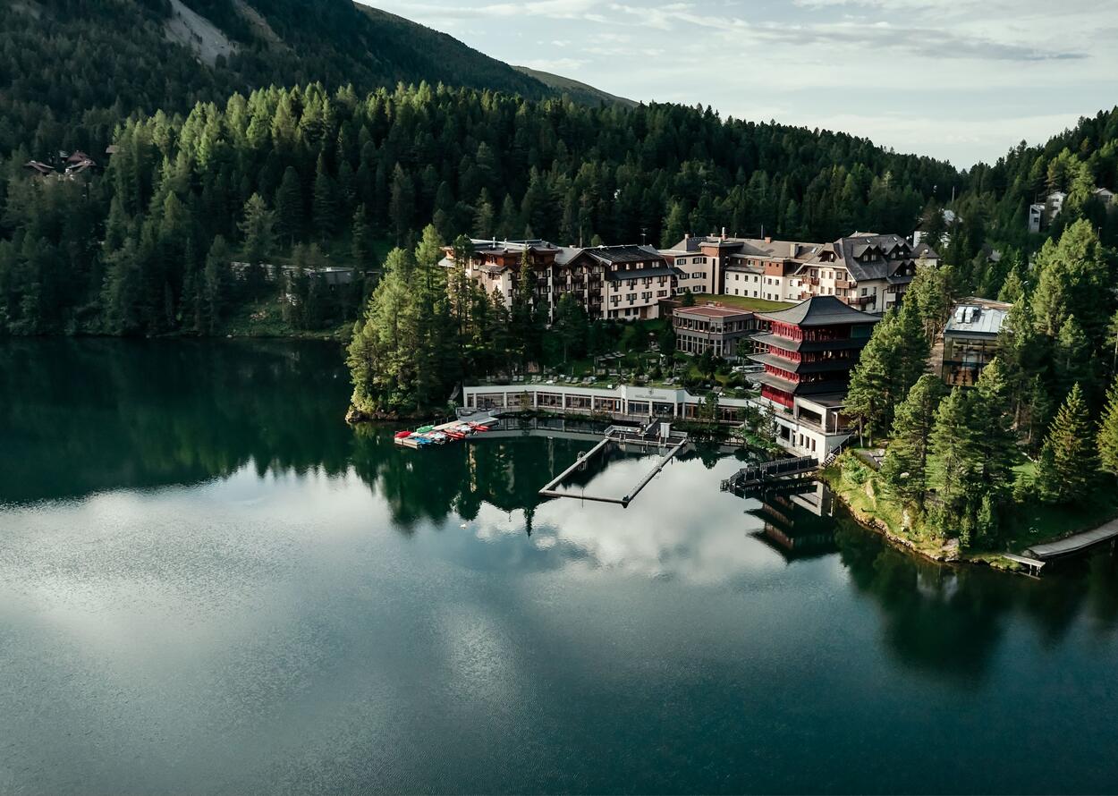 A hotel resort is idyllically situated by a calm lake, surrounded by dense forest and an impressive mountain landscape.