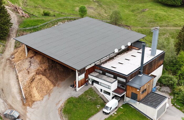Luftaufnahme eines modernen Gebäudekomplexes in ländlicher Umgebung. Neben dem Gebäude liegen große Holzspäne- oder Hackschnitzelhaufen, was auf ein Biomasse- oder Heizwerk hinweist. Rundherum sind grüne Wiesen, Wege und einzelne Bäume zu sehen.