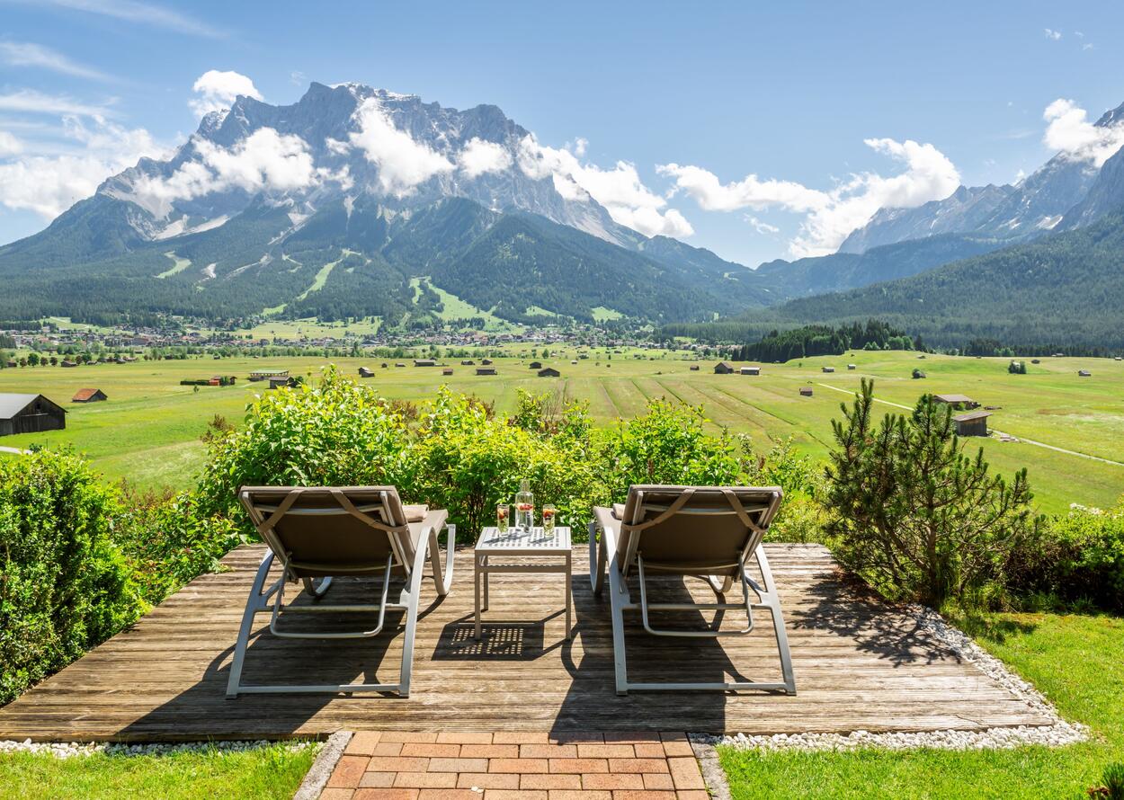 Zwei Liegestühle stehen vor einer beeindruckenden Berglandschaft mit grünen Wiesen und blauem Himmel.