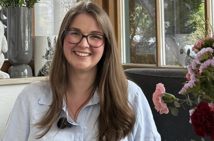 Porträt einer lächelnden Frau mit Brille in einem lichtdurchfluteten Innenraum mit Blumenarrangements.