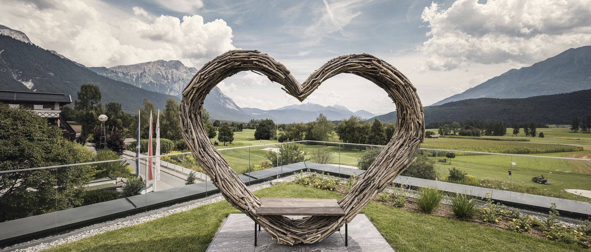 Herzförmige Holzskulptur mit Sitzbank vor alpiner Landschaft und Bergpanorama.