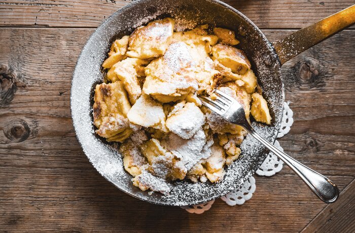 Pfanne mit Kaiserschmarrn, bestäubt mit Puderzucker, auf einem rustikalen Holztisch. Daneben steht ein kleines Schälchen mit Apfelmus.