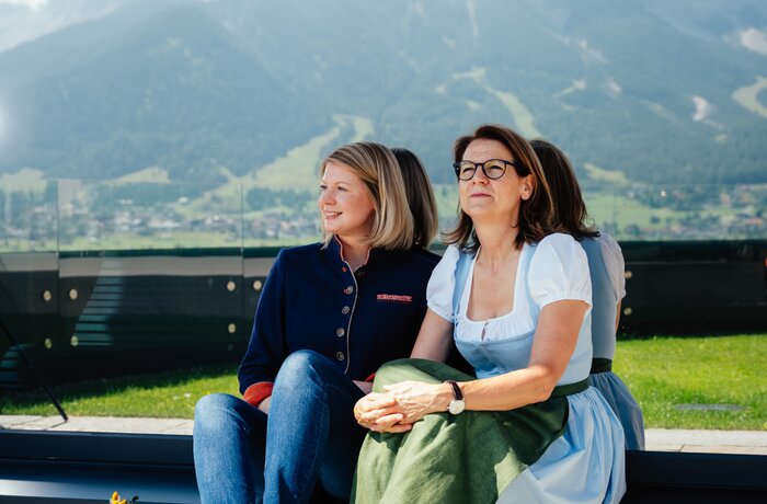 Two women are sitting outside in front of an impressive mountain landscape, enjoying the view on a sunny day.