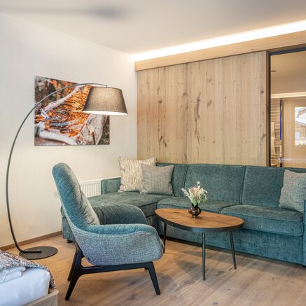 A cozy hotel room with a modern sofa, armchair, and warm wooden elements. A floor lamp and subtle decoration create a relaxing atmosphere.