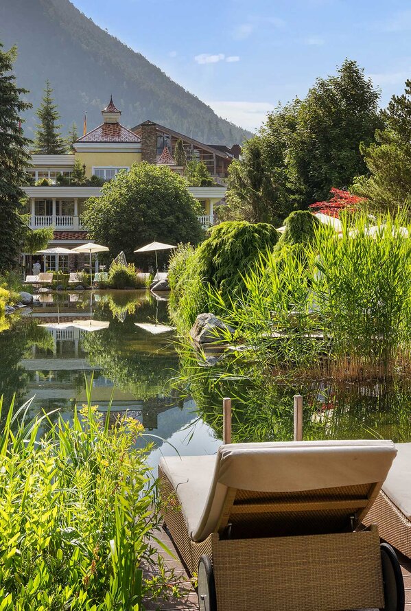 Naturbadeteich mit Sonnenliegen, Sonnenschirmen und Gartenanlage vor Bergkulisse