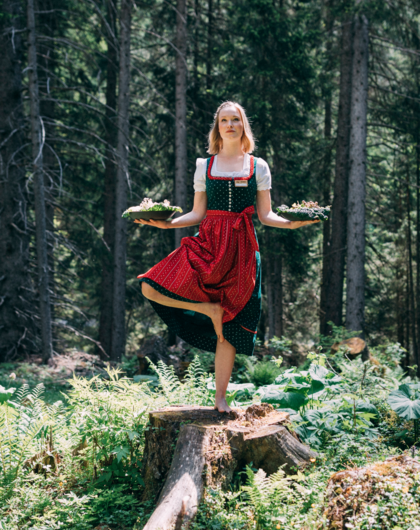 Frau im Dirndl balanciert auf Baumstumpf mit Tabletts im Wald