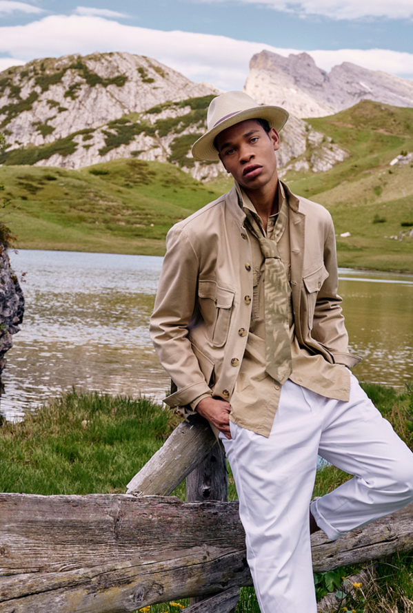 Mann mit Hut in beiger Jacke und weißer Hose vor Berglandschaft mit See