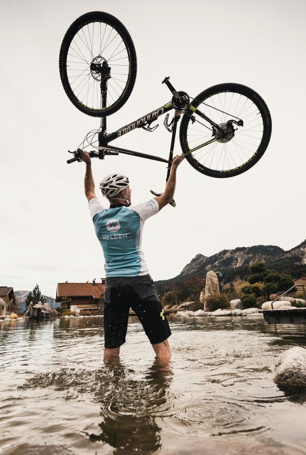 Radfahrer steht im Wasser und hält Mountainbike hoch