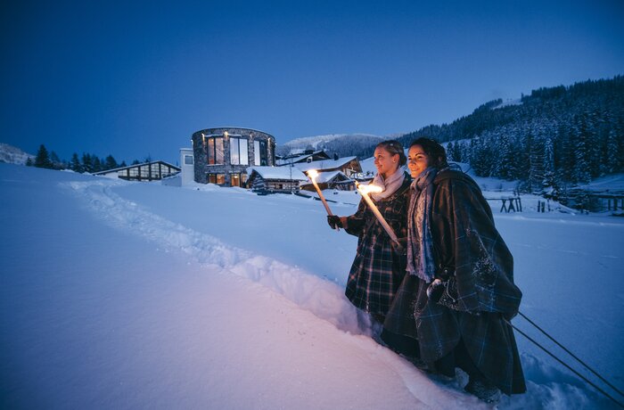 Zwei Frauen mit Fackeln im Schnee vor beleuchtetem Gebäude in verschneiter Landschaft
