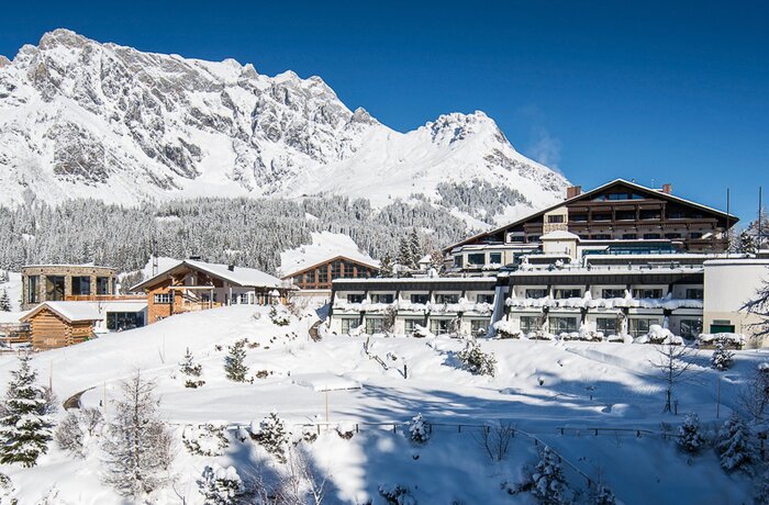 Verschneite Häuser und Hotelanlage vor schneebedecktem Bergmassiv unter blauem Himmel