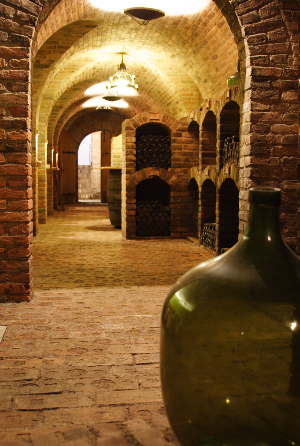 Gewölbekeller mit Ziegelsteinen, Weinregalen und großer grüner Flasche im Vordergrund