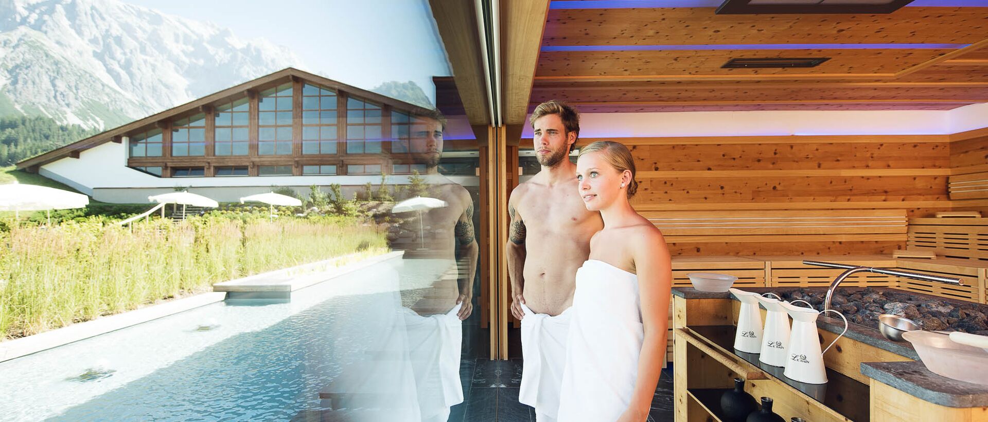 Couple in towels in front of window with pool and mountain panorama in wellness area