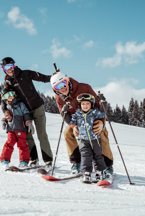 Familie mit zwei Kindern beim Skifahren auf einer schneebedeckten Piste vor bewaldetem Hintergrund