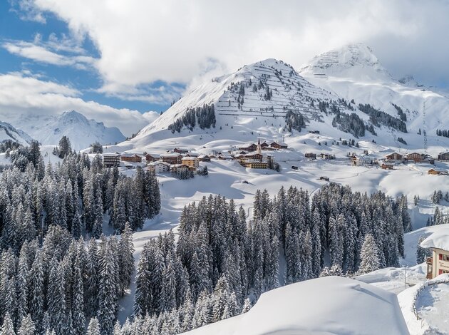 Winterlandschaft mit schneebedeckten Bergen, Tannen und Häusern unter blauem Himmel mit Wolken
