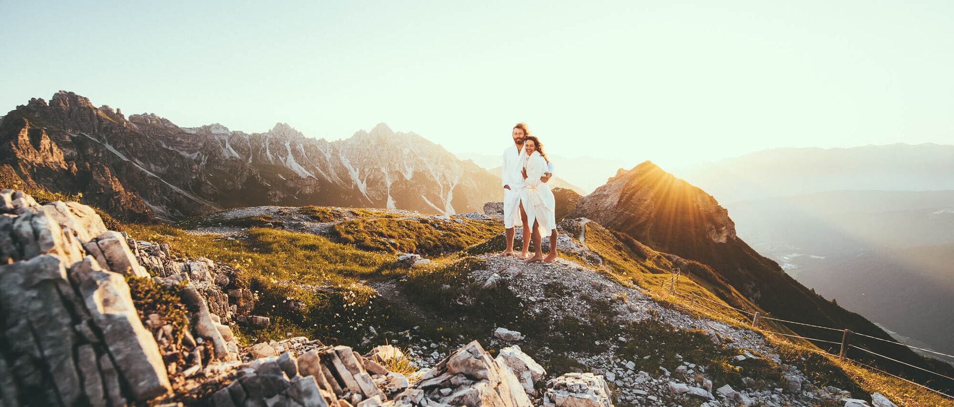 Couple in white bathrobes standing barefoot on a rock in the mountains at sunrise