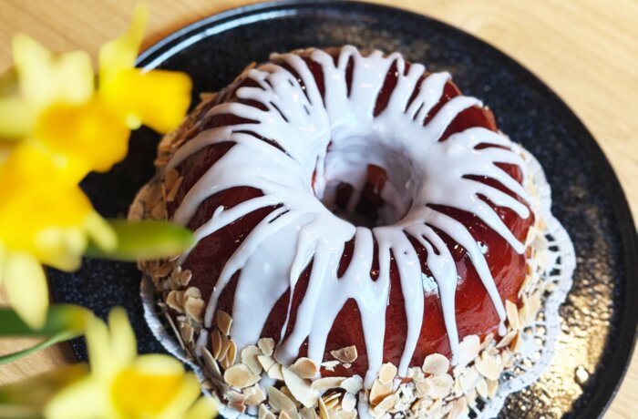 Gugelhupf mit Zuckerguss und Mandeln auf schwarzem Teller, Narzissen im Vordergrund