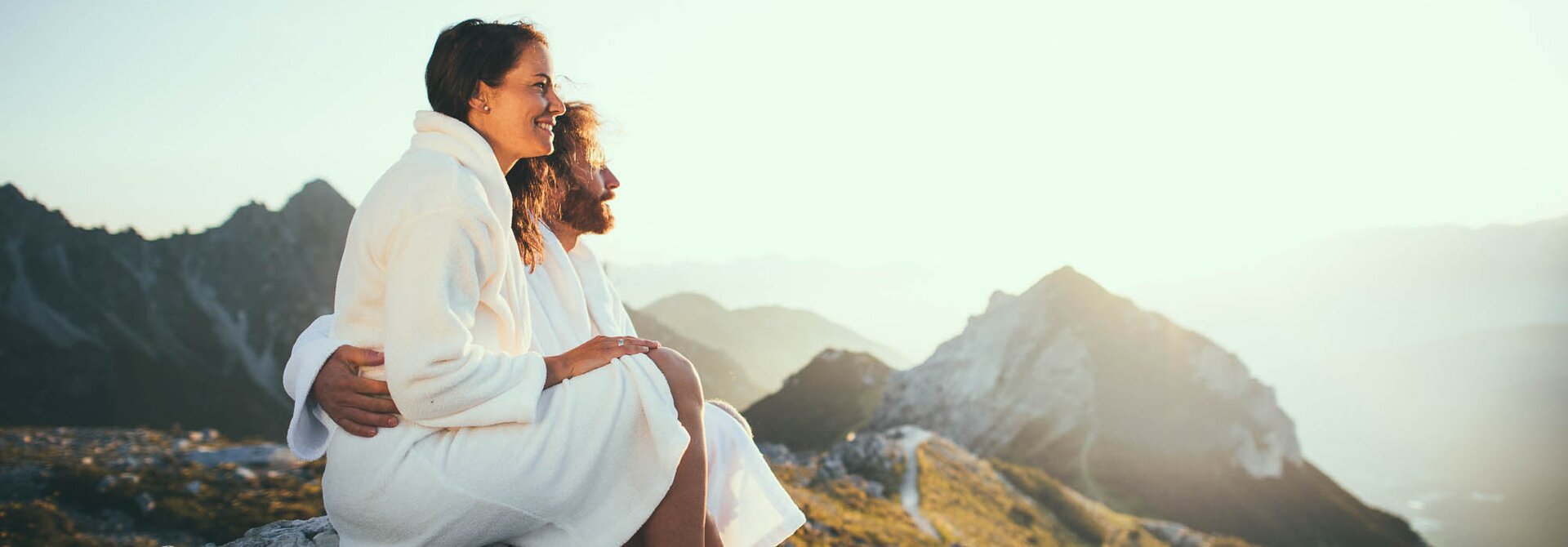 Paar in Bademänteln sitzt auf Felsen und blickt auf Berglandschaft