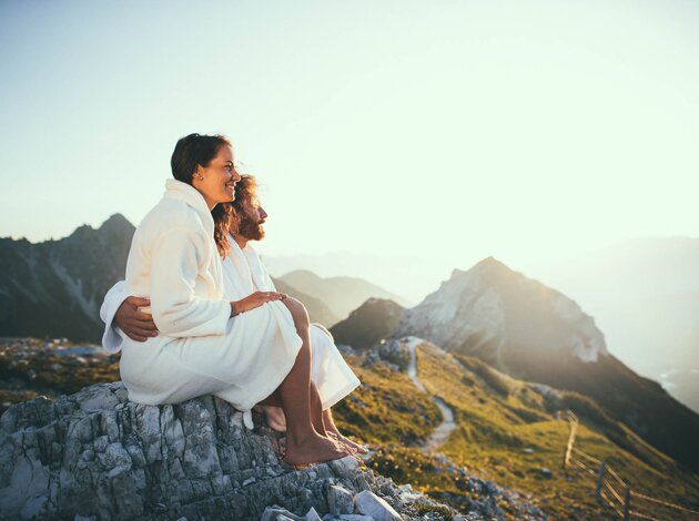 Paar in Bademänteln sitzt auf Felsen und blickt auf Berglandschaft