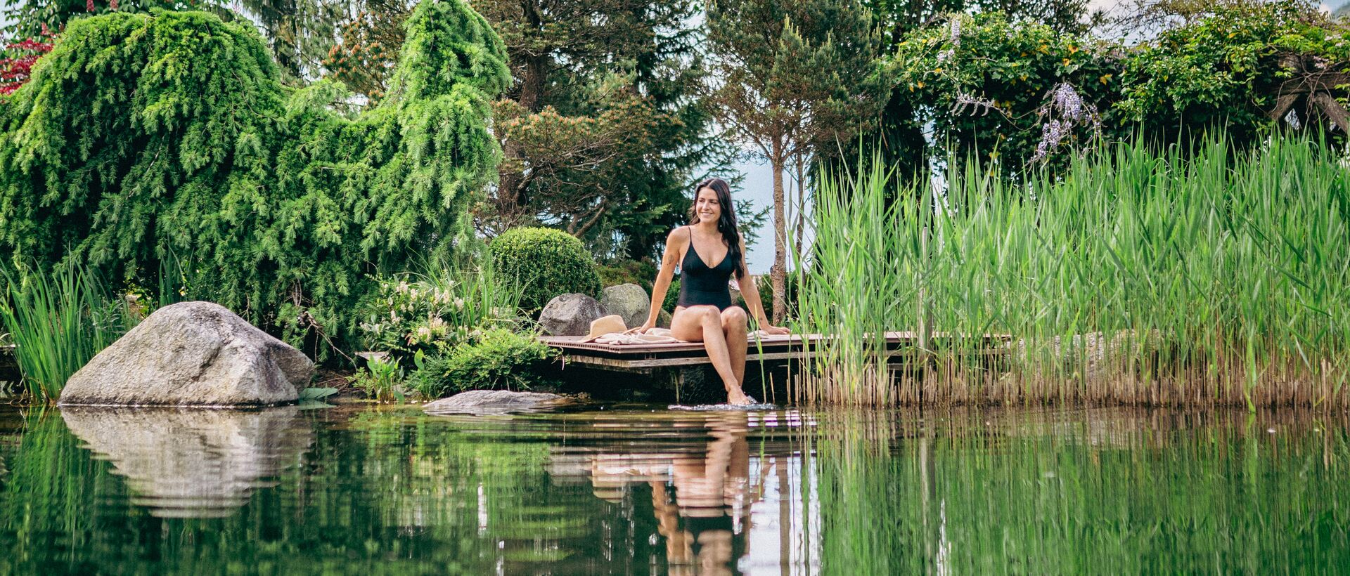 Frau sitzt barfuß auf Holzsteg am Teich, umgeben von üppiger Vegetation und Spiegelungen im Wasser