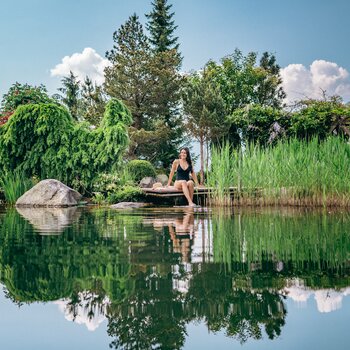 Woman sitting barefoot on wooden jetty by pond, surrounded by lush vegetation and reflections in the water