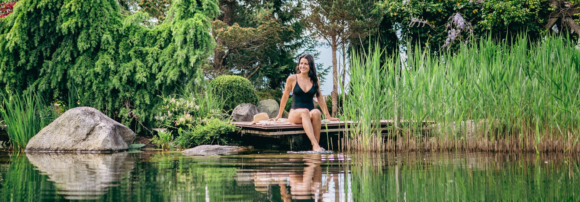 Woman sitting barefoot on wooden jetty by pond, surrounded by lush vegetation and reflections in the water