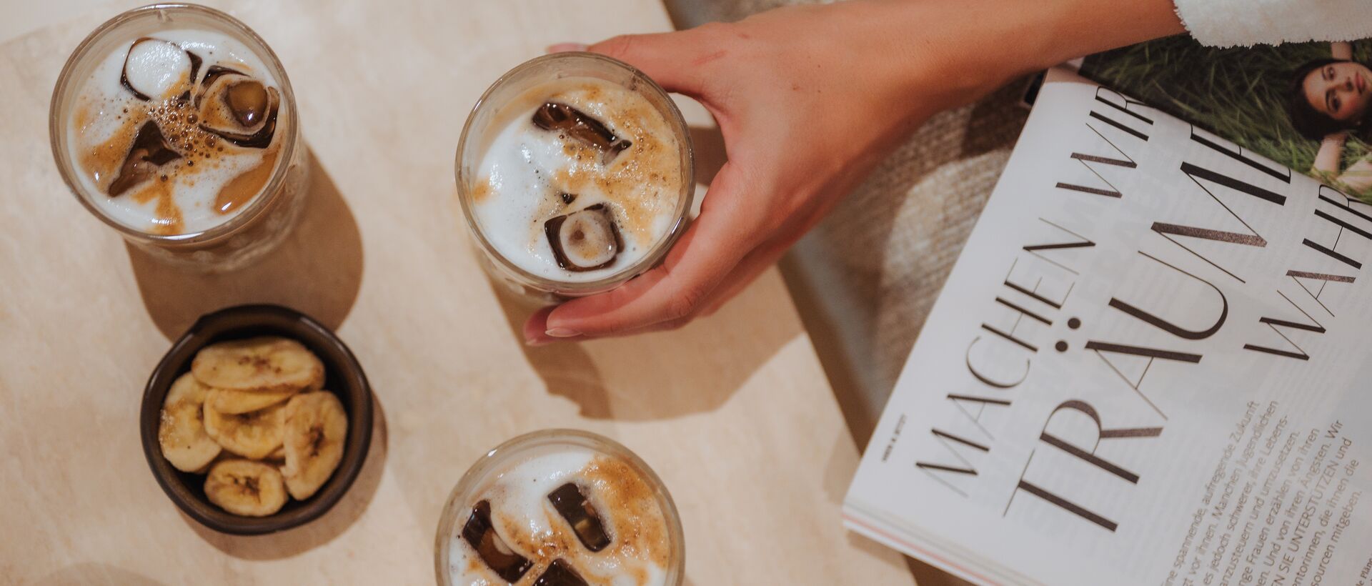 Three drinks with ice cubes, banana chips and a hand on a table with a magazine