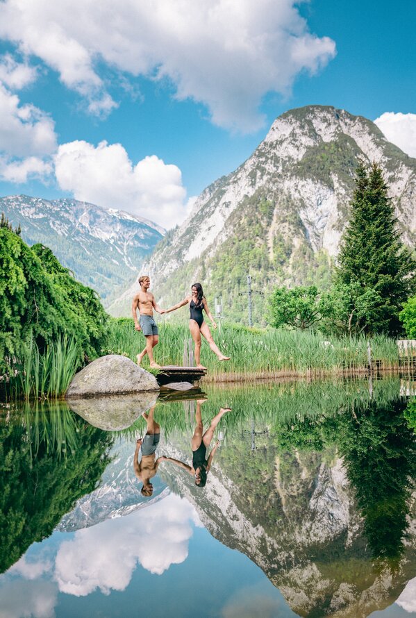Paar in Badekleidung balanciert auf Holzsteg über Bergsee, Spiegelung im Wasser