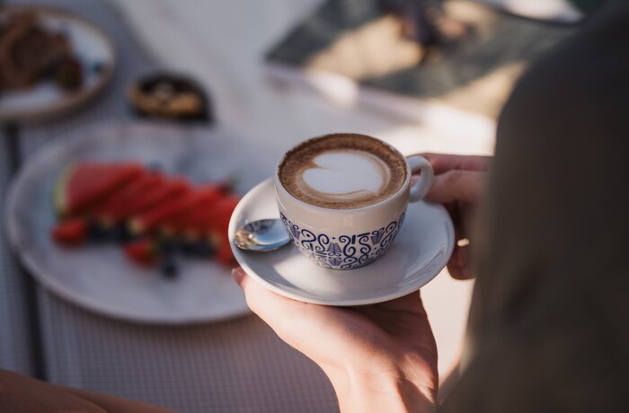 Frau hält Tasse Cappuccino mit Milchschaumherz, Teller mit Erdbeeren und Gebäck im Hintergrund