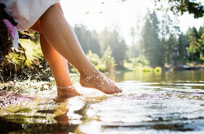 Eine Person taucht ihre Füße in einen See am Waldrand. Sonnenlicht spiegelt sich im Wasser, während kleine Wasserspritzer entstehen.