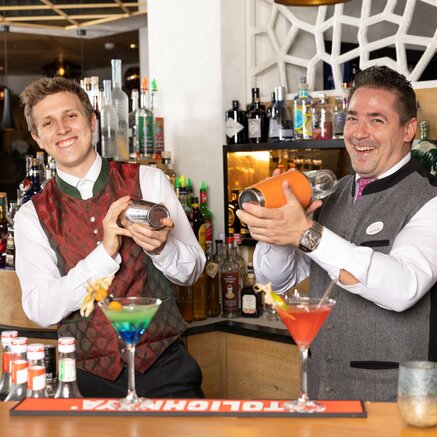Two bartenders mix cocktails behind a bar with colorful drinks and spirits
