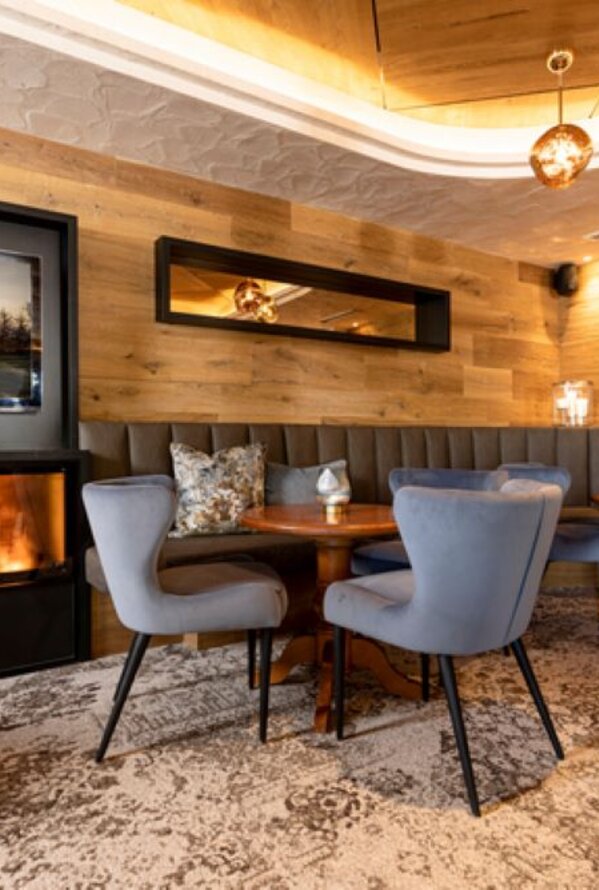 Gemütliche Lounge mit Kamin, Holzwand, blauen Stühlen und runden Tischen auf gemustertem Teppich