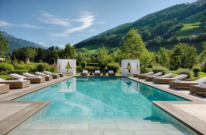 Türkisblauer Pool vor grüner Berglandschaft mit Liegen und weißen Pavillons