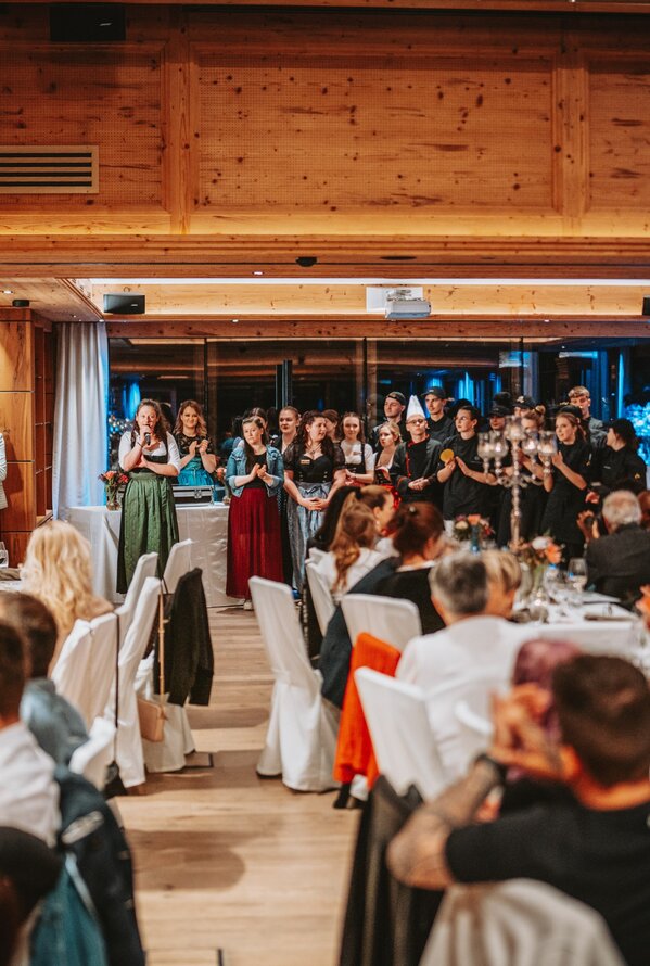 Gruppe in Tracht singt vor Publikum in festlichem Saal mit gedeckten Tischen