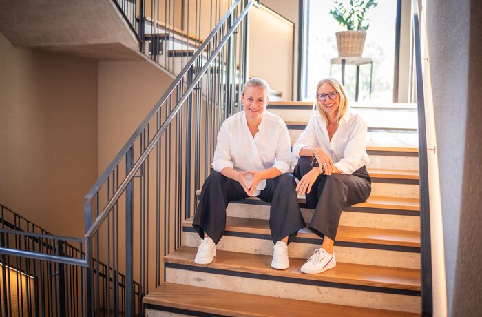 Two women sit relaxed on a modern staircase, smiling at the camera – stylish, friendly, and authentic.