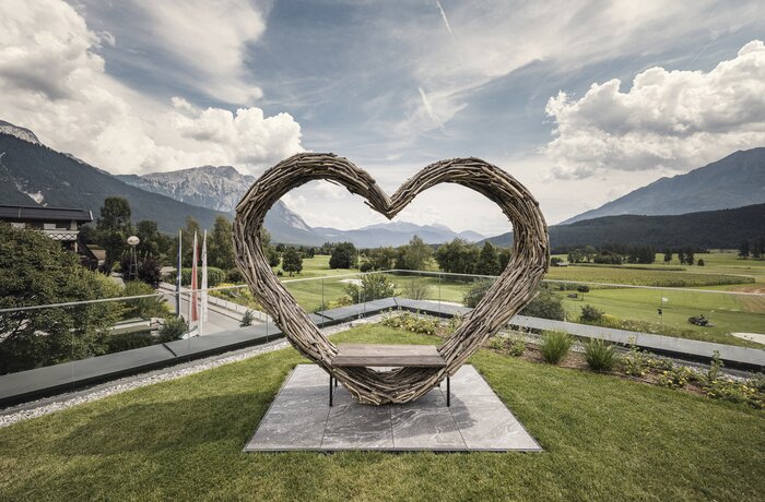 Herzförmige Holzskulptur mit Sitzbank vor alpiner Landschaft und Bergpanorama.