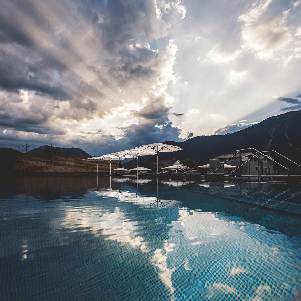 Pool mit Sonnenschirmen vor Bergkulisse unter bewölktem Himmel