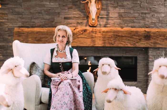 Frau Gürtler im Dirndl sitzt auf einem Sessel neben zwei Stoffschafen vor einem Kamin