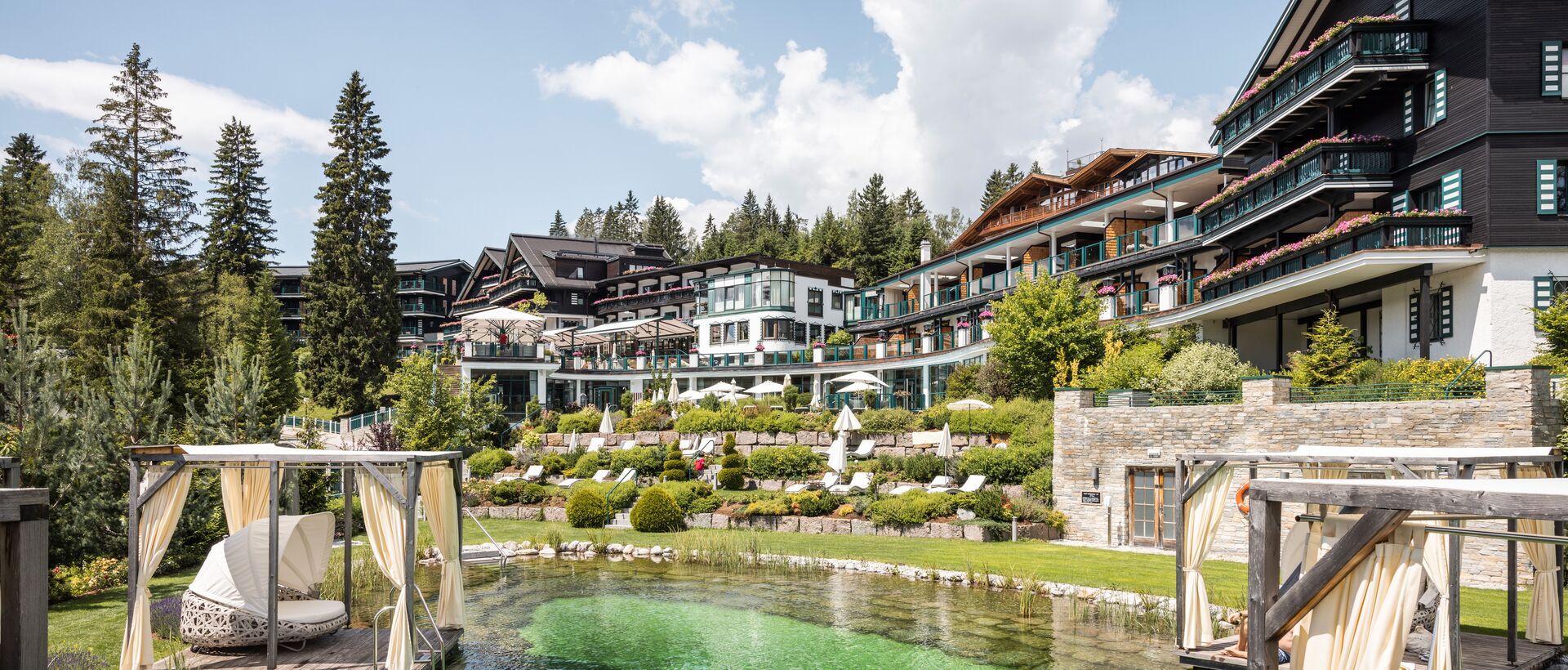 Großes Wellnesshotel mit Gartenanlage, Naturbadeteich und privaten Ruhebereichen inmitten von Bäumen und Bergen.