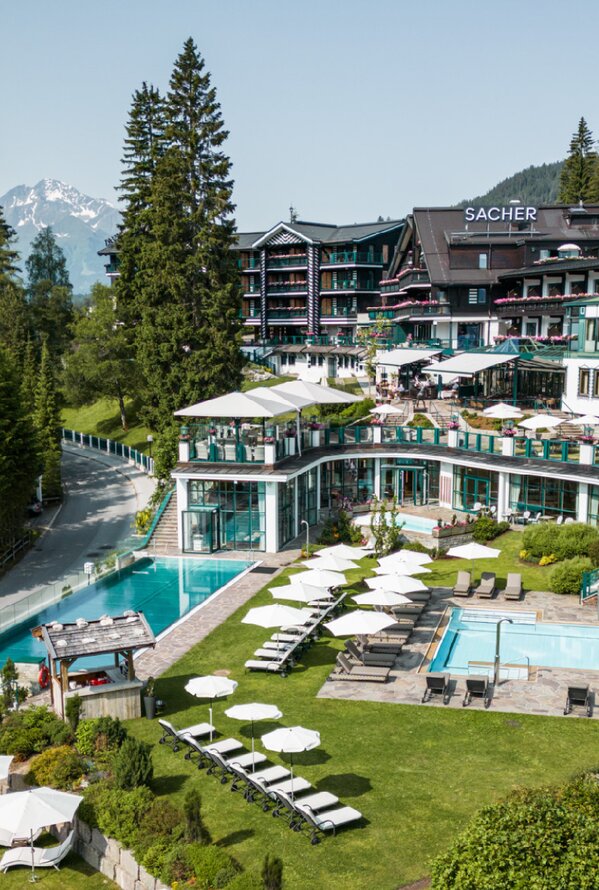 Aerial view of a wellness hotel with outdoor pools, sunbathing lawns, and landscaped gardens surrounded by tall trees and mountains.