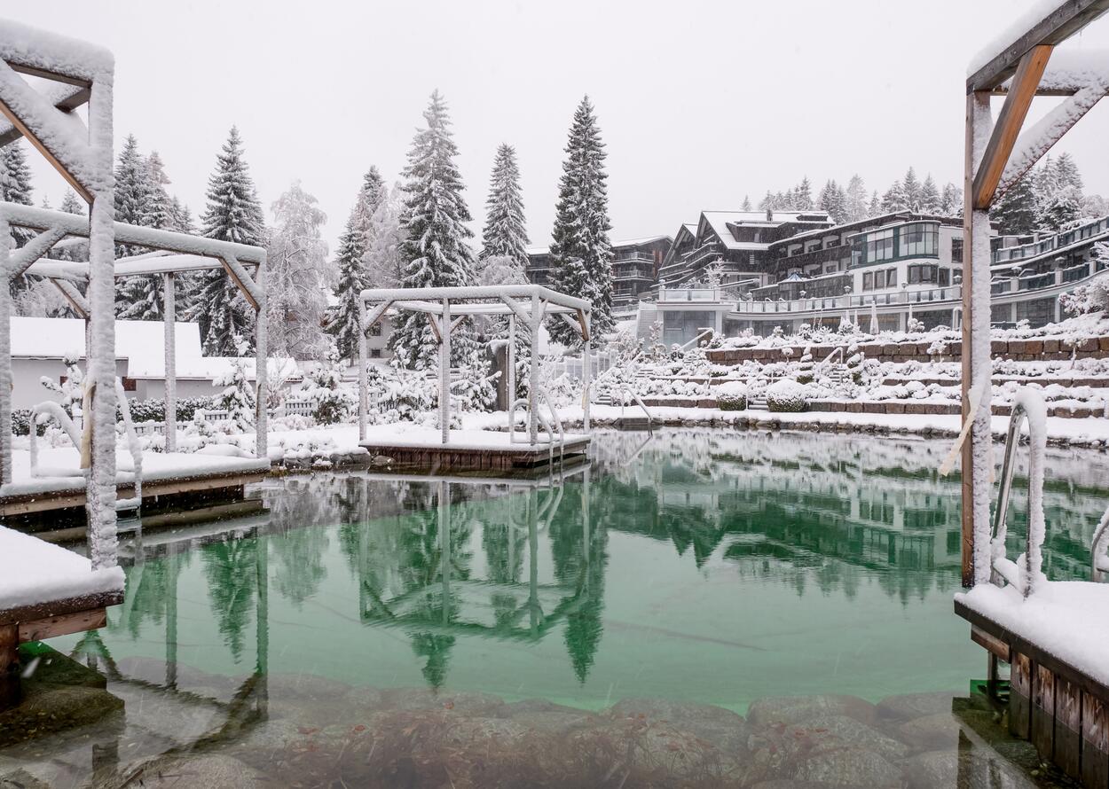 Winter natural bathing lake with snow-covered trees and buildings, reflection in the clear, greenish water