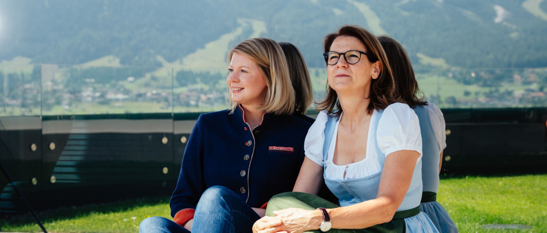 Zwei Frauen sitzen draußen vor einer beeindruckenden Berglandschaft und genießen die Aussicht bei sonnigem Wetter.