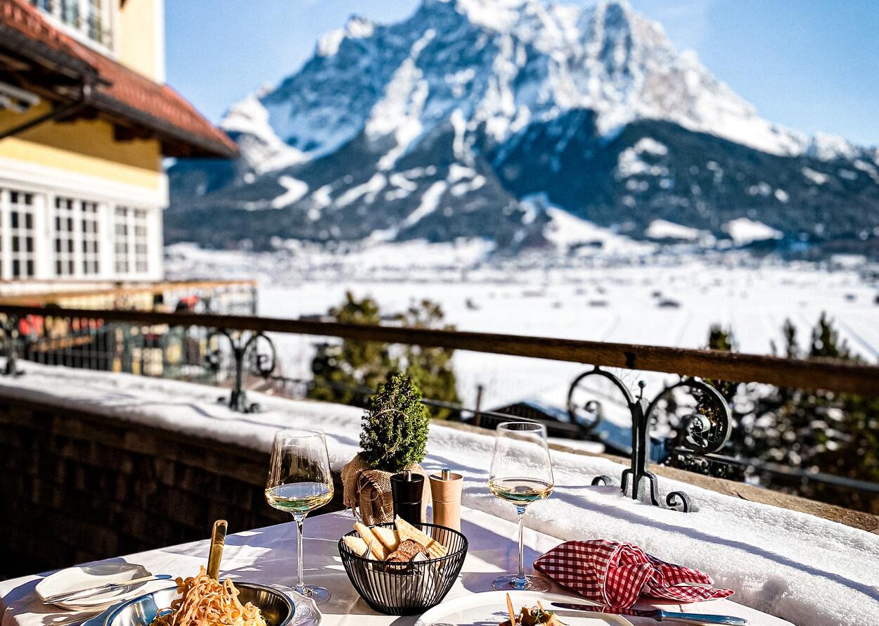 Ein stilvoll gedeckter Tisch auf einer Sonnenterrasse mit Blick auf verschneite Berge und winterliche Landschaft.