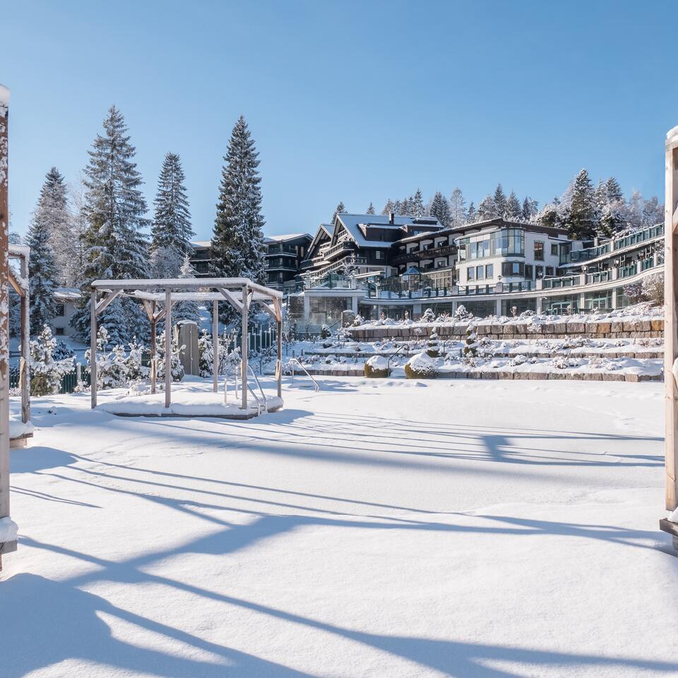 Verschneiter Naturbadesee im Winter mit Hotelanlage und Tannenbäumen unter blauem Himmel