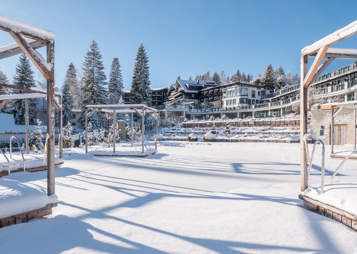 Verschneiter Naturbadesee im Winter mit Hotelanlage und Tannenbäumen unter blauem Himmel