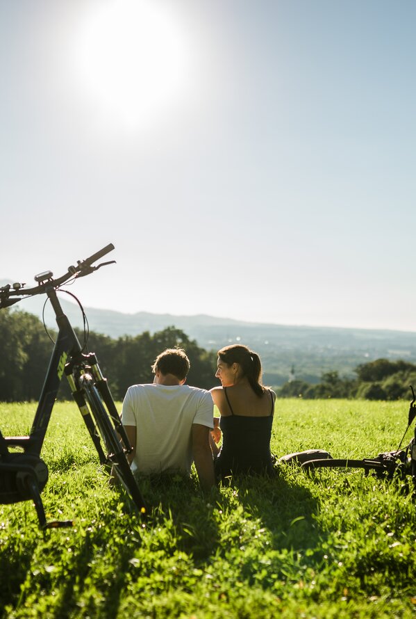 Paar sitzt auf Wiese mit Fahrrädern und blickt in die Ferne unter strahlender Sonne
