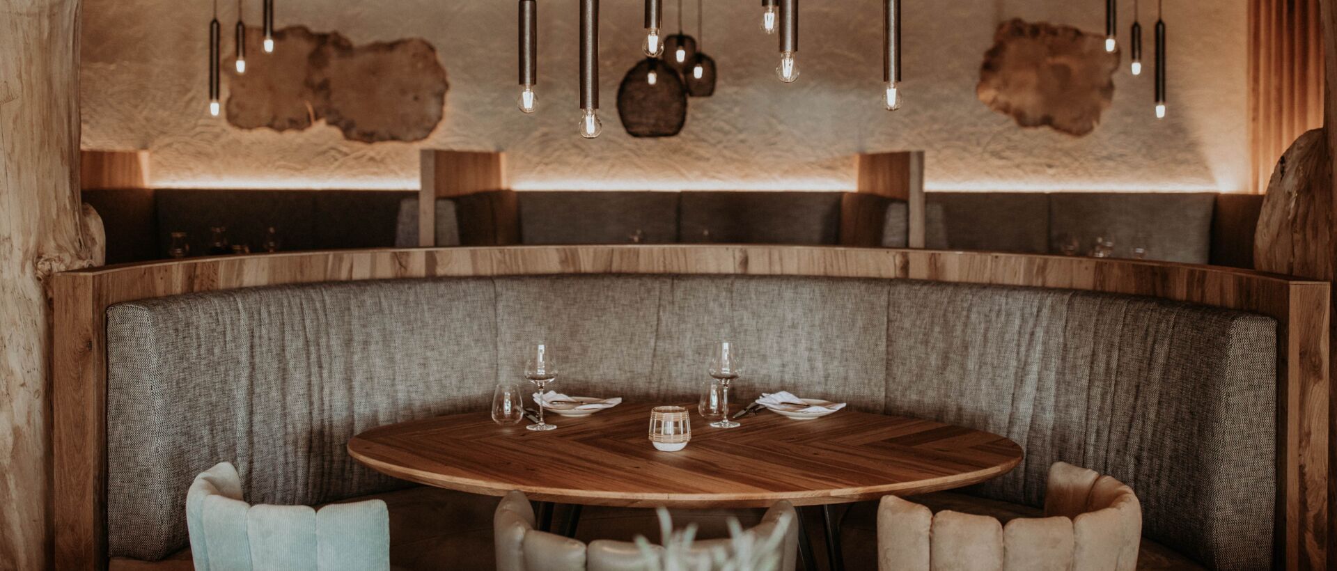 Restaurant mit runder Holztischgruppe, grauer Sitzbank und hängenden Röhrenleuchten vor holzvertäfelter Wand