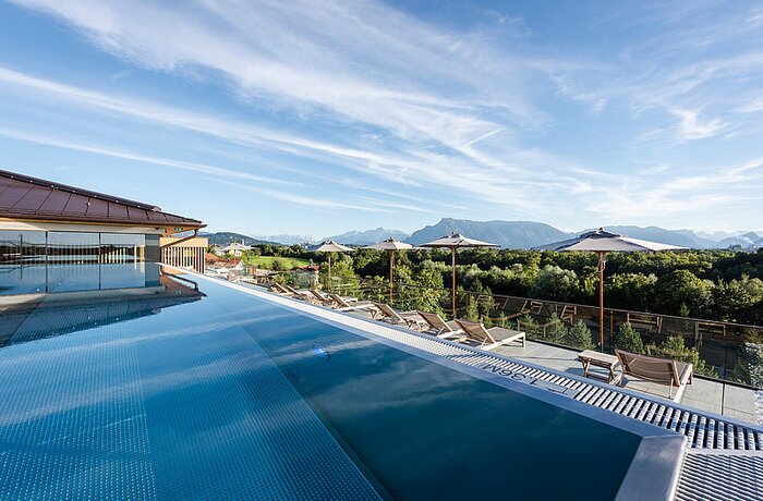 Infinity-Pool mit Liegen und Sonnenschirmen vor grüner Landschaft und Bergen unter blauem Himmel