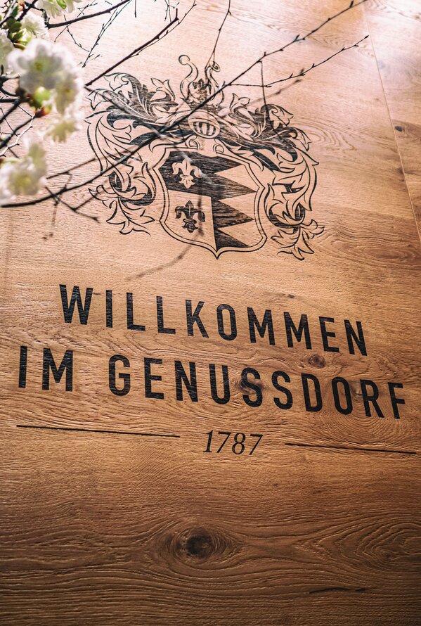Holzwand mit Wappen und dem Schriftzug "Willkommen im Genussdorf 1787"